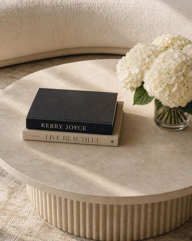 Coffee table design study seen from above, fluted travertine table with stacked books and white hydrangeas beside a cream curved sofa.