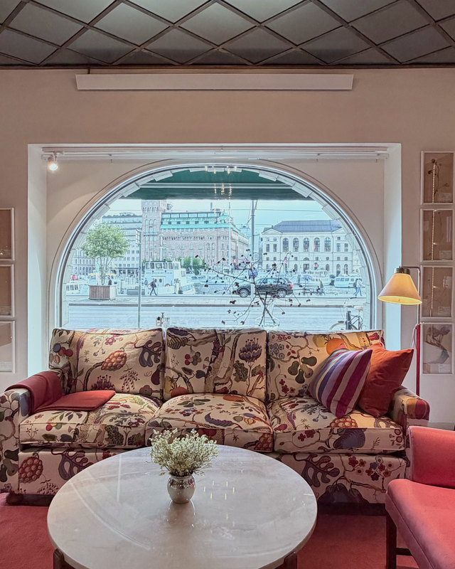 A sofa in a large floral print under an arched window looking out over the water in Stockholm, with a round marble table and a red carpet.
