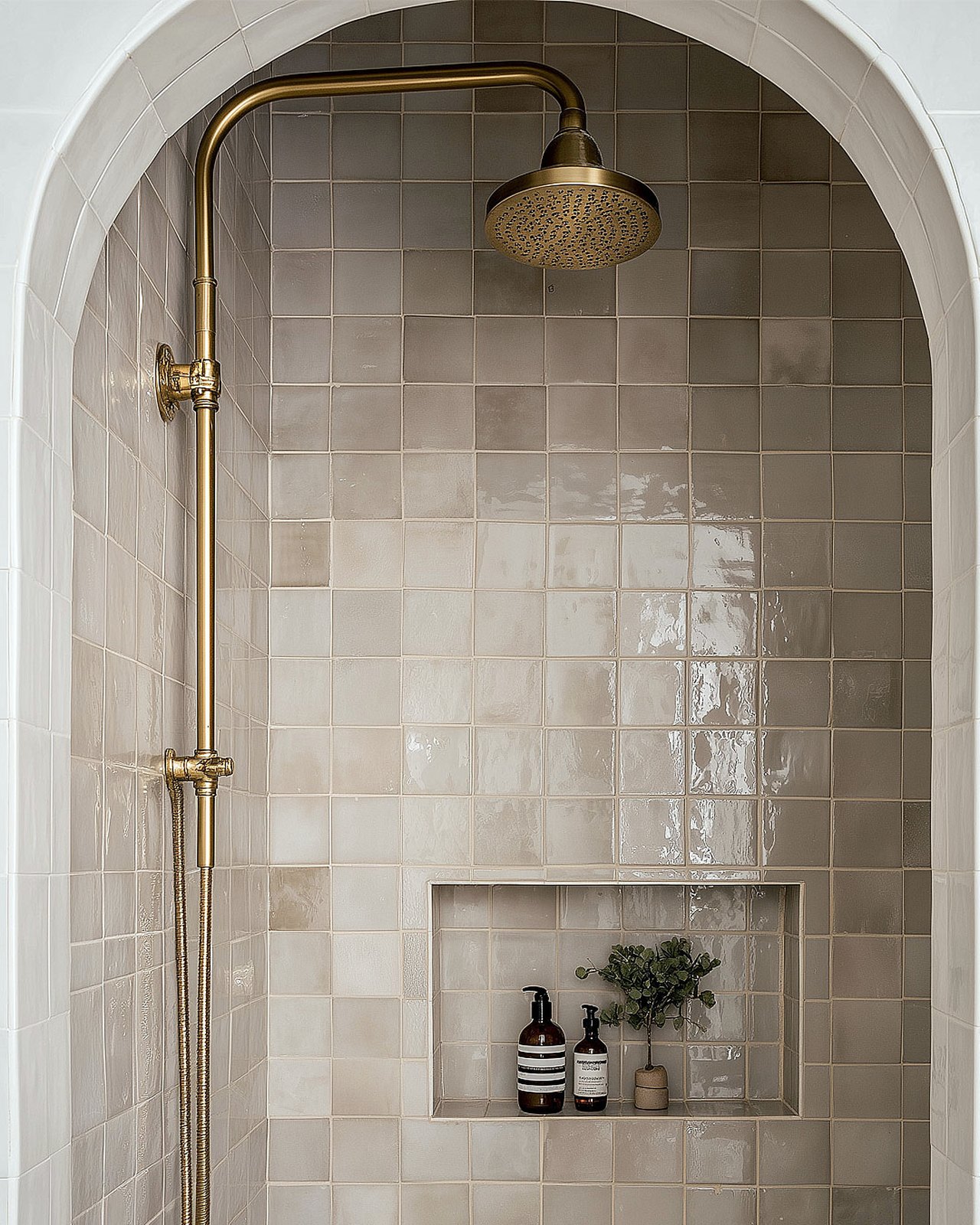 An arched shower niche in glazed tile, brass rain shower and a small shelf of bottles and a eucalyptus sprig, Studio Sakaia, Dubai