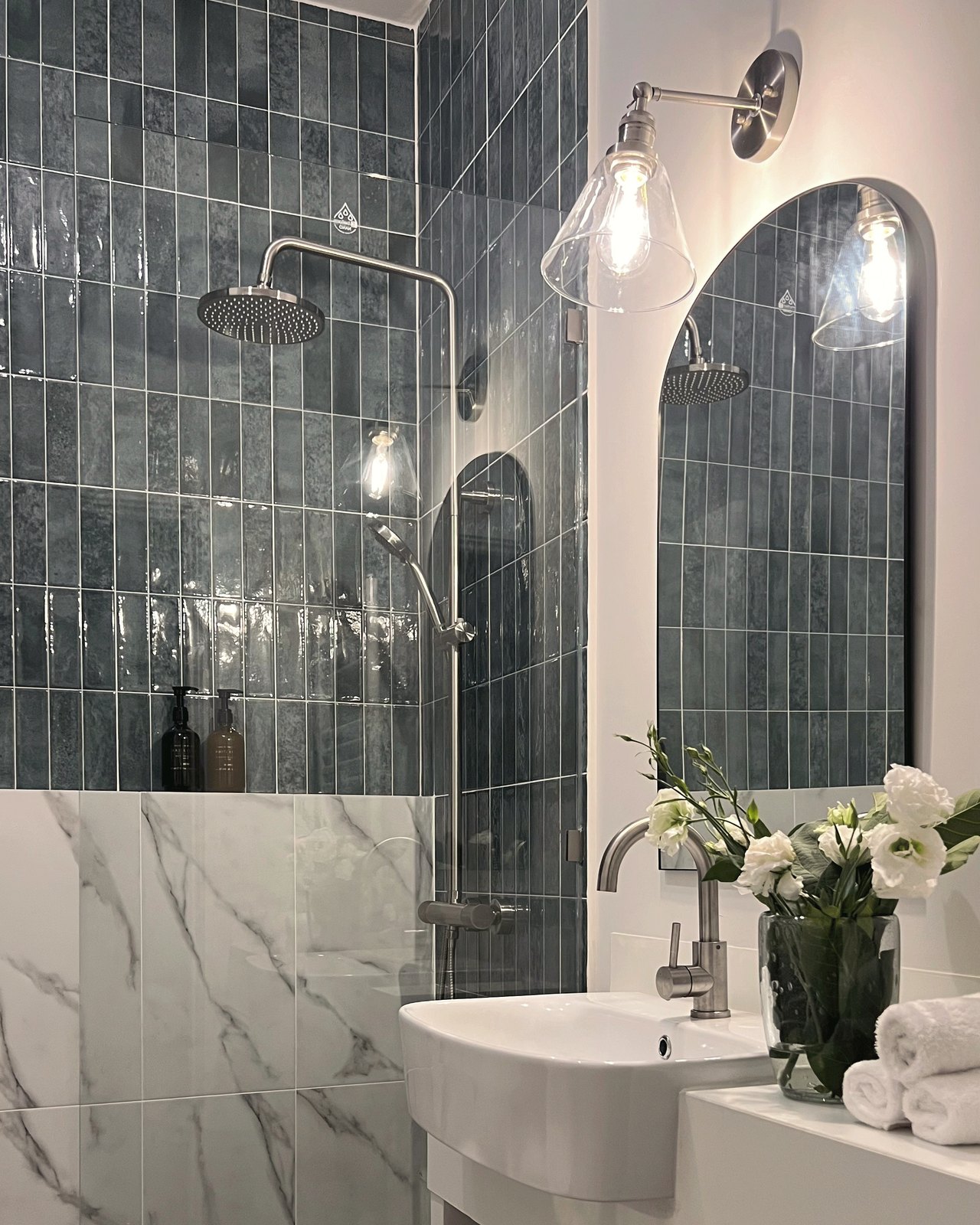 A vanity corner with an arched mirror above a wall-hung basin, dark tile and fresh cut flowers, Studio Sakaia, Dubai