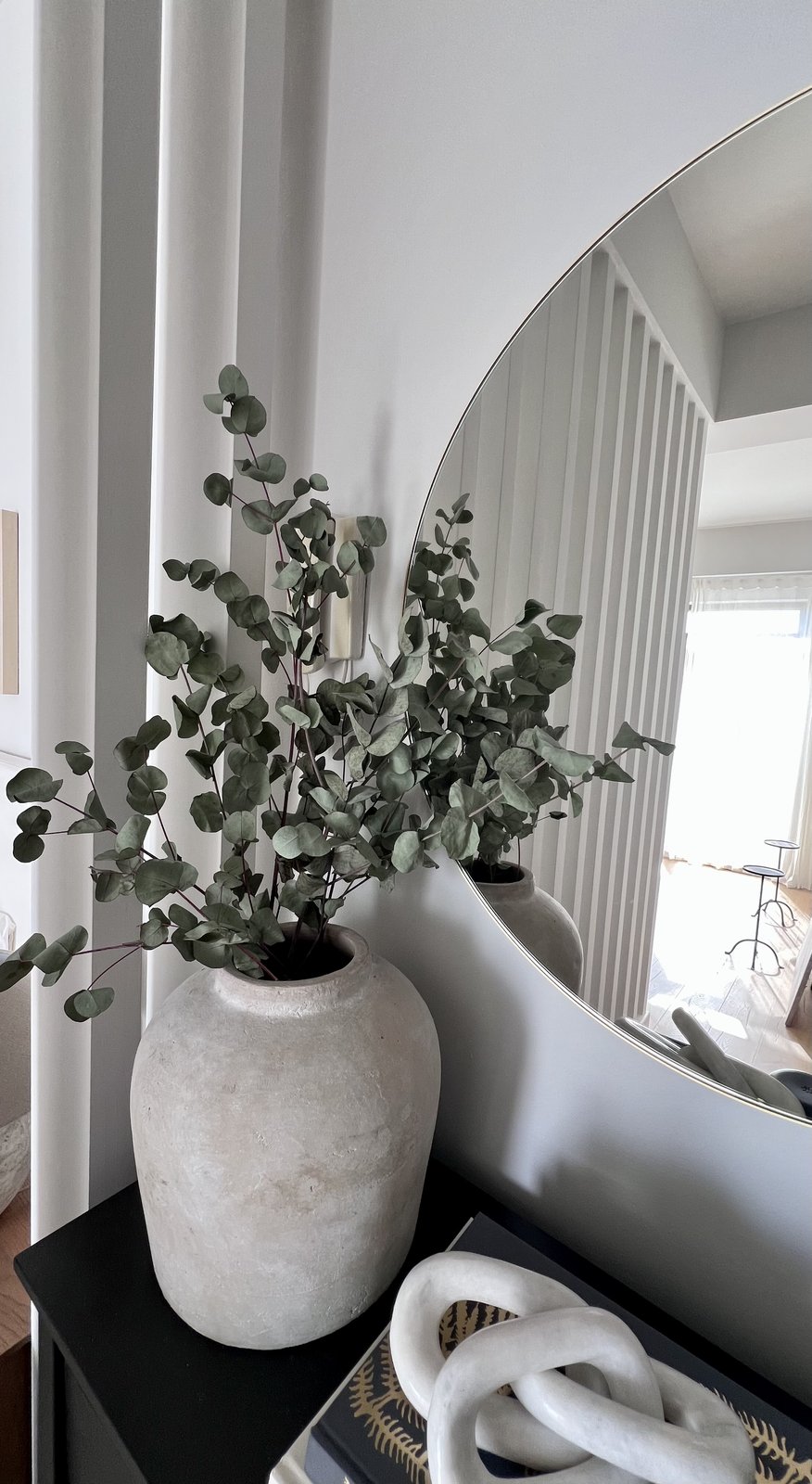 A styled entry console with a round brass mirror, eucalyptus in a stone vase, stacked design books and a candle, the slatted room divider reflected in the mirror, Studio Sakaia's own Dubai home
