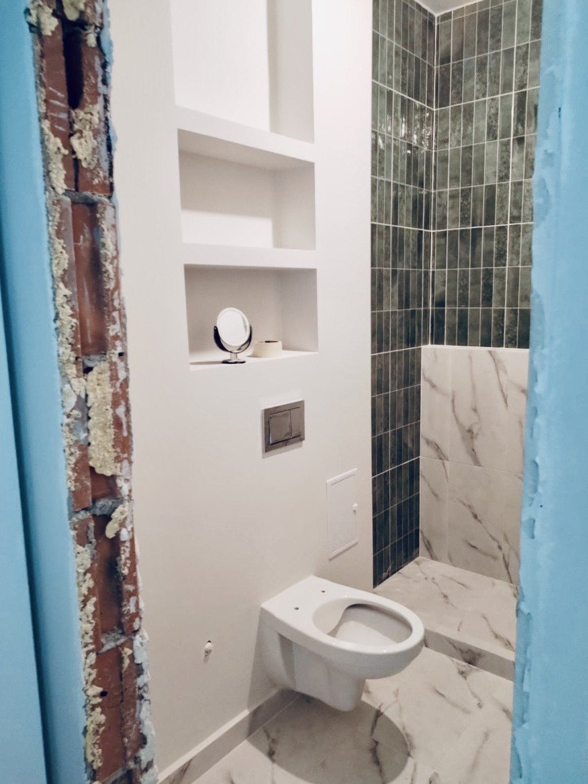 The finished niche: white shelves, vertical green tile and marble, with the waterproofing membrane still visible at the door reveal, Studio Sakaia