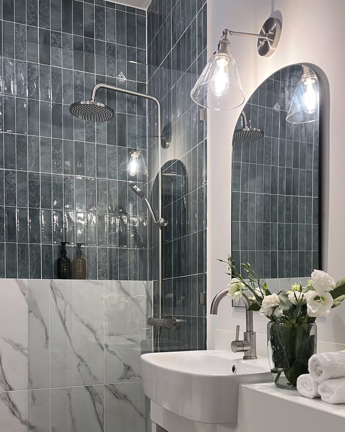 A styled bathroom corner with an arched mirror, wall light, floating basin and fresh flowers against the fluted teal tile, Studio Sakaia, Dubai