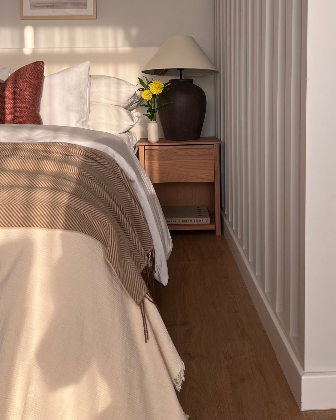 Morning light on a styled bedroom nightstand with oak table, dark ceramic lamp, and fresh flowers.