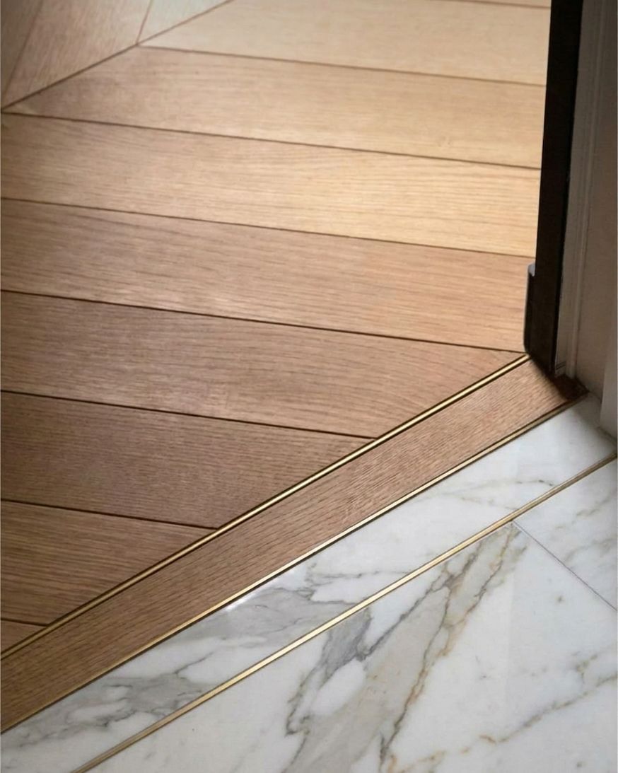Brass inlay threshold where chevron oak flooring meets veined marble at a doorway.
