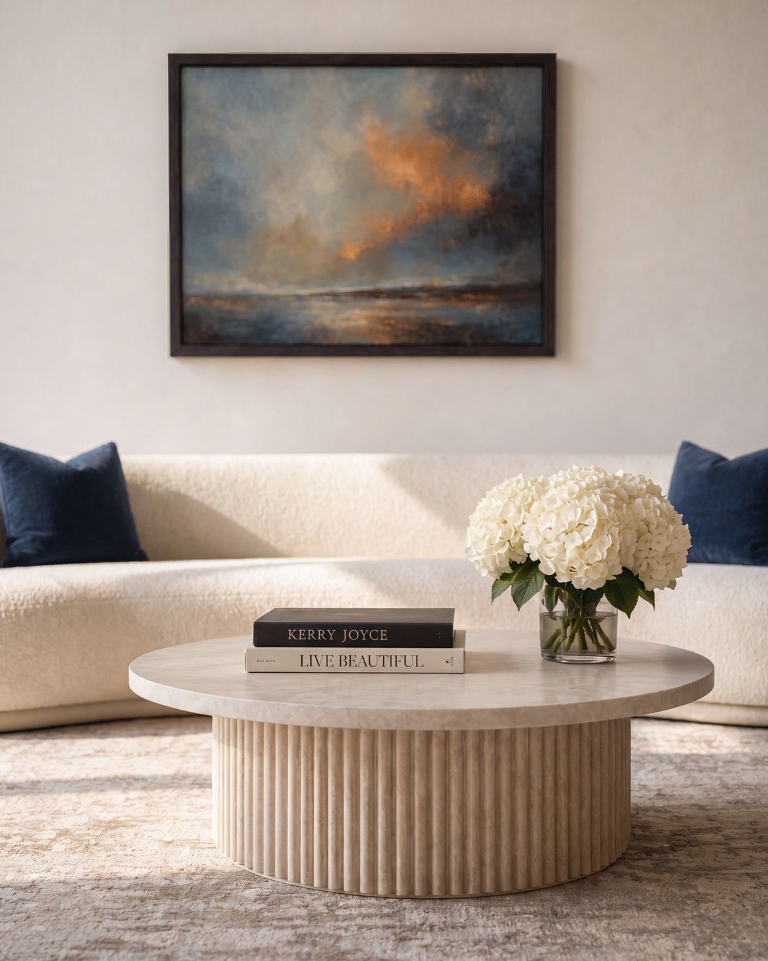 Coffee table vignette, fluted travertine table with stacked books and white hydrangeas in front of a cream curved sofa.