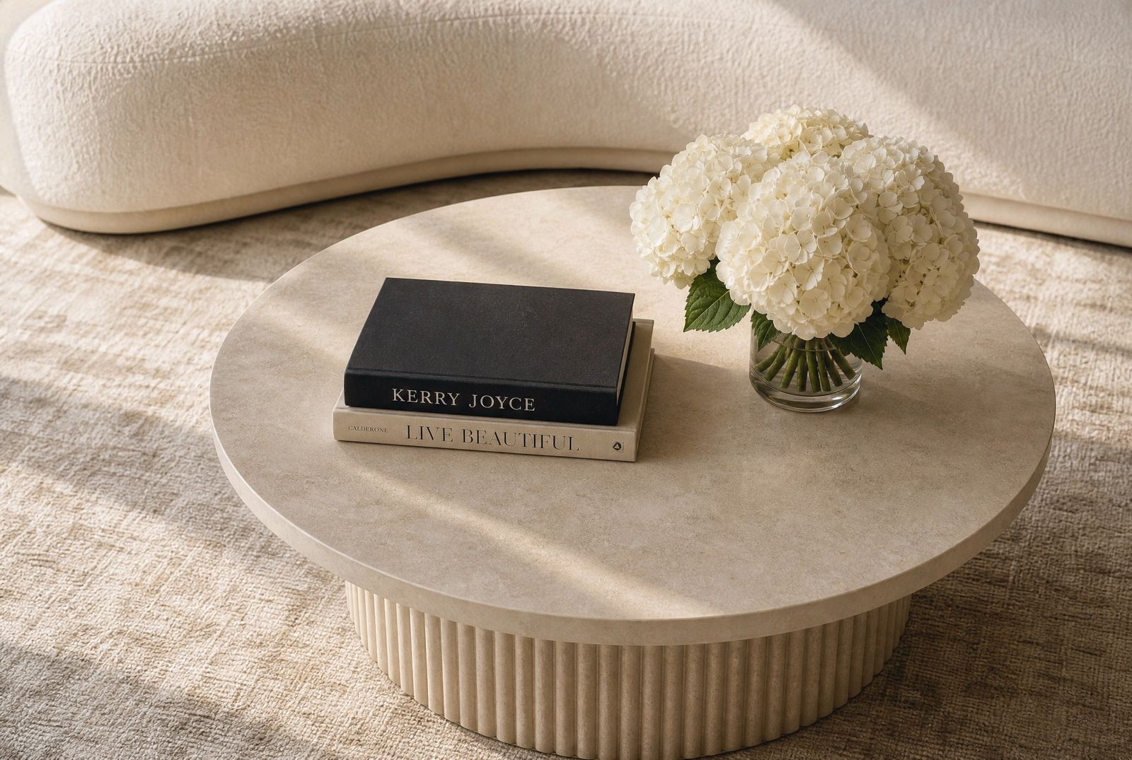 Coffee table design study seen from above, fluted travertine table with stacked books and white hydrangeas beside a cream curved sofa.