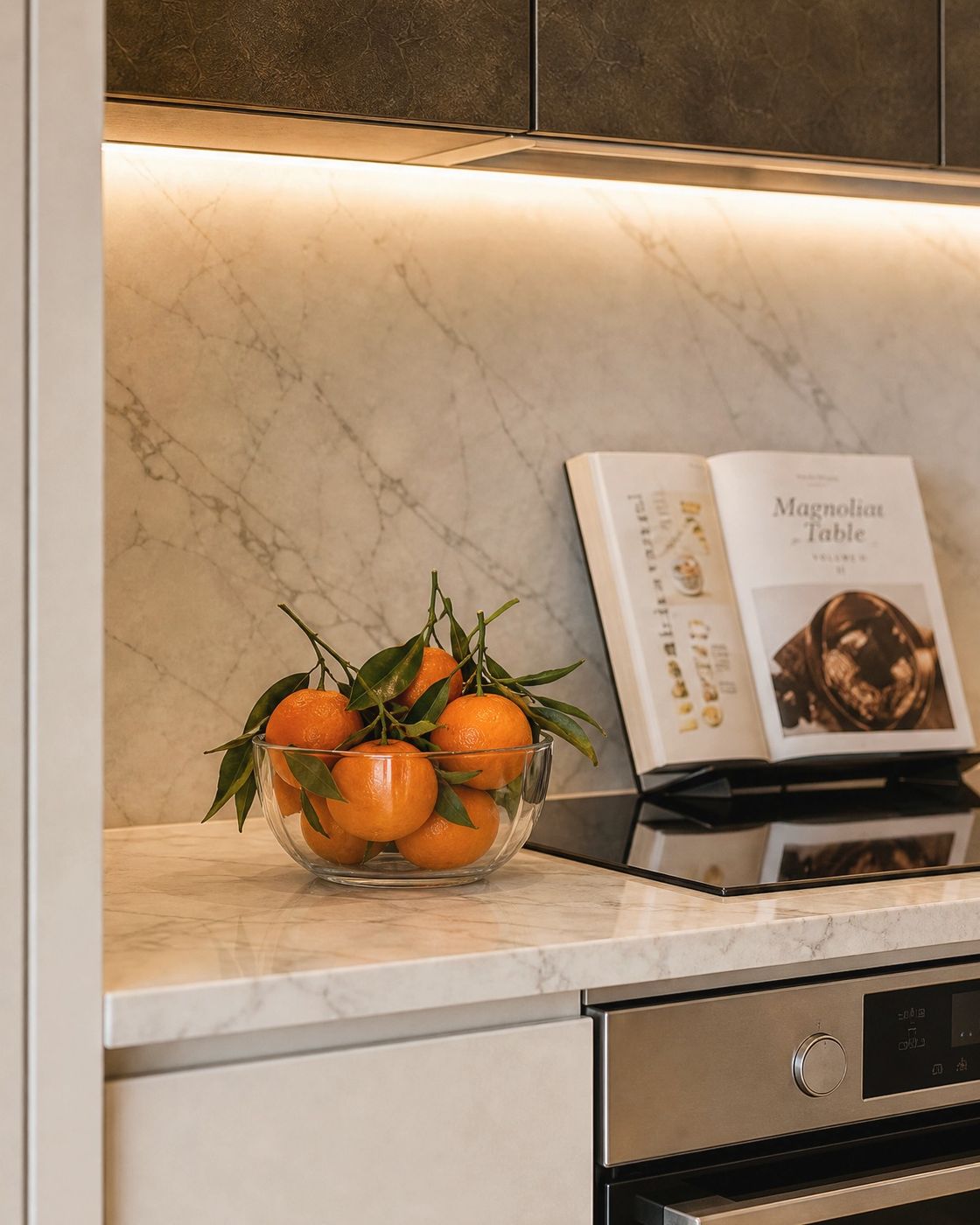 Kitchen counter vignette, clear glass bowl of clementines with the leaves on and an open cookbook against a marble backsplash.