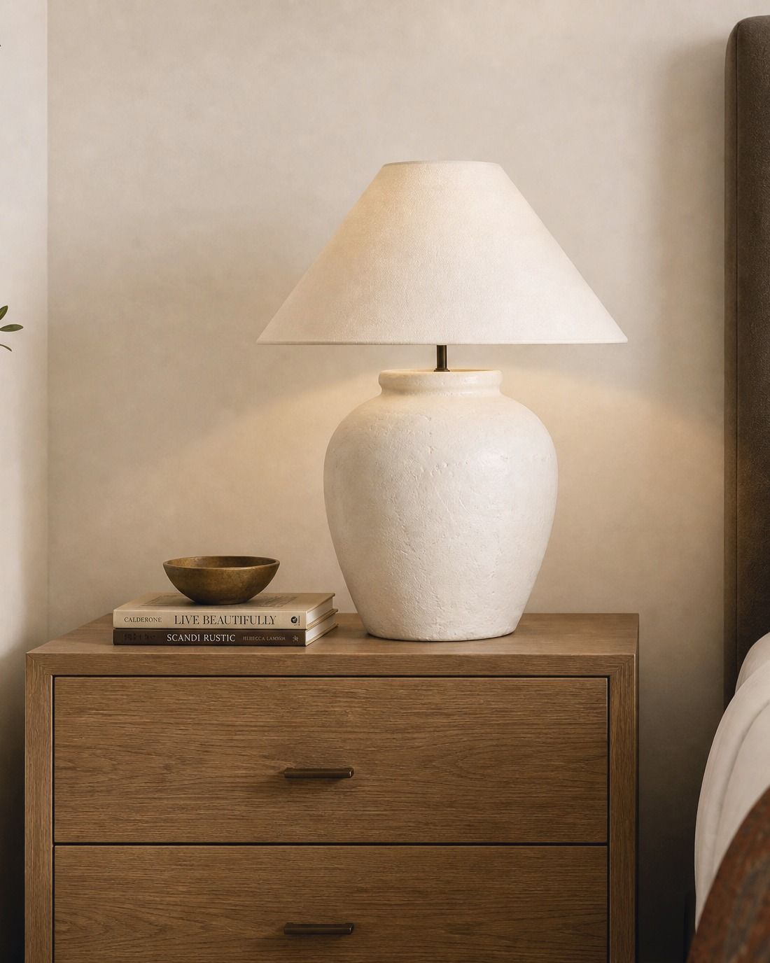 Nightstand vignette, ivory ceramic lamp, small brass bowl and two books on an oak nightstand beside a bed.