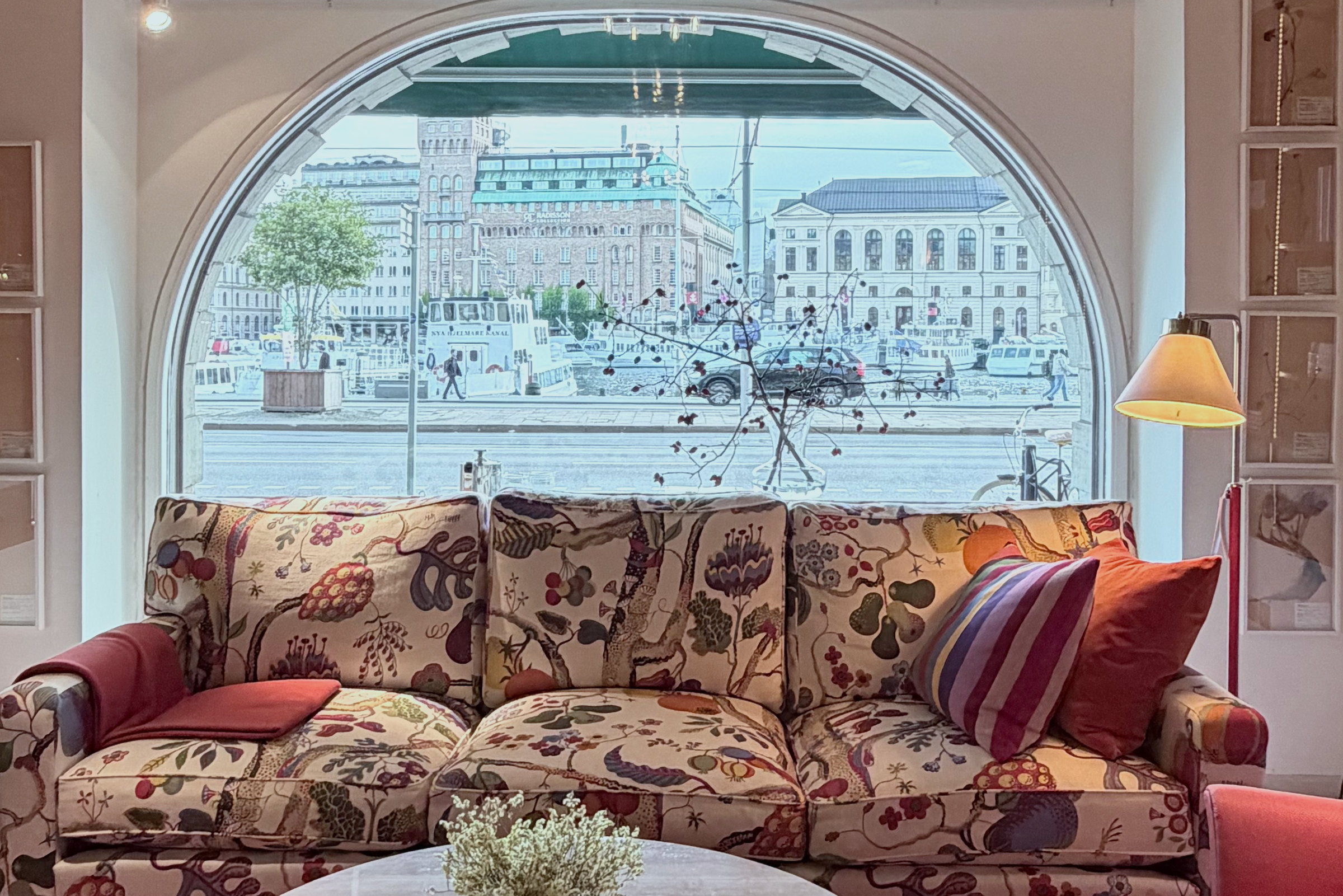 A sofa in a large floral print under an arched window looking out over the water in Stockholm, with a round marble table and a red carpet.