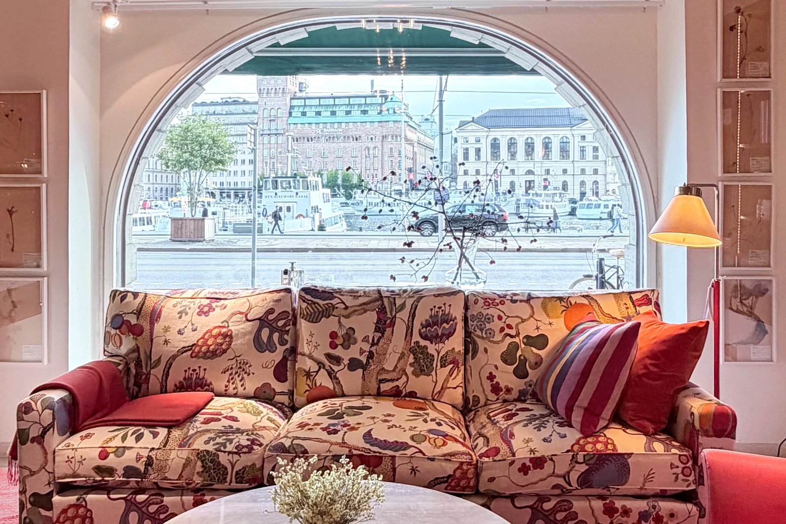 A sofa in a large floral print under an arched window looking out over the water in Stockholm, with a round marble table and a red carpet.