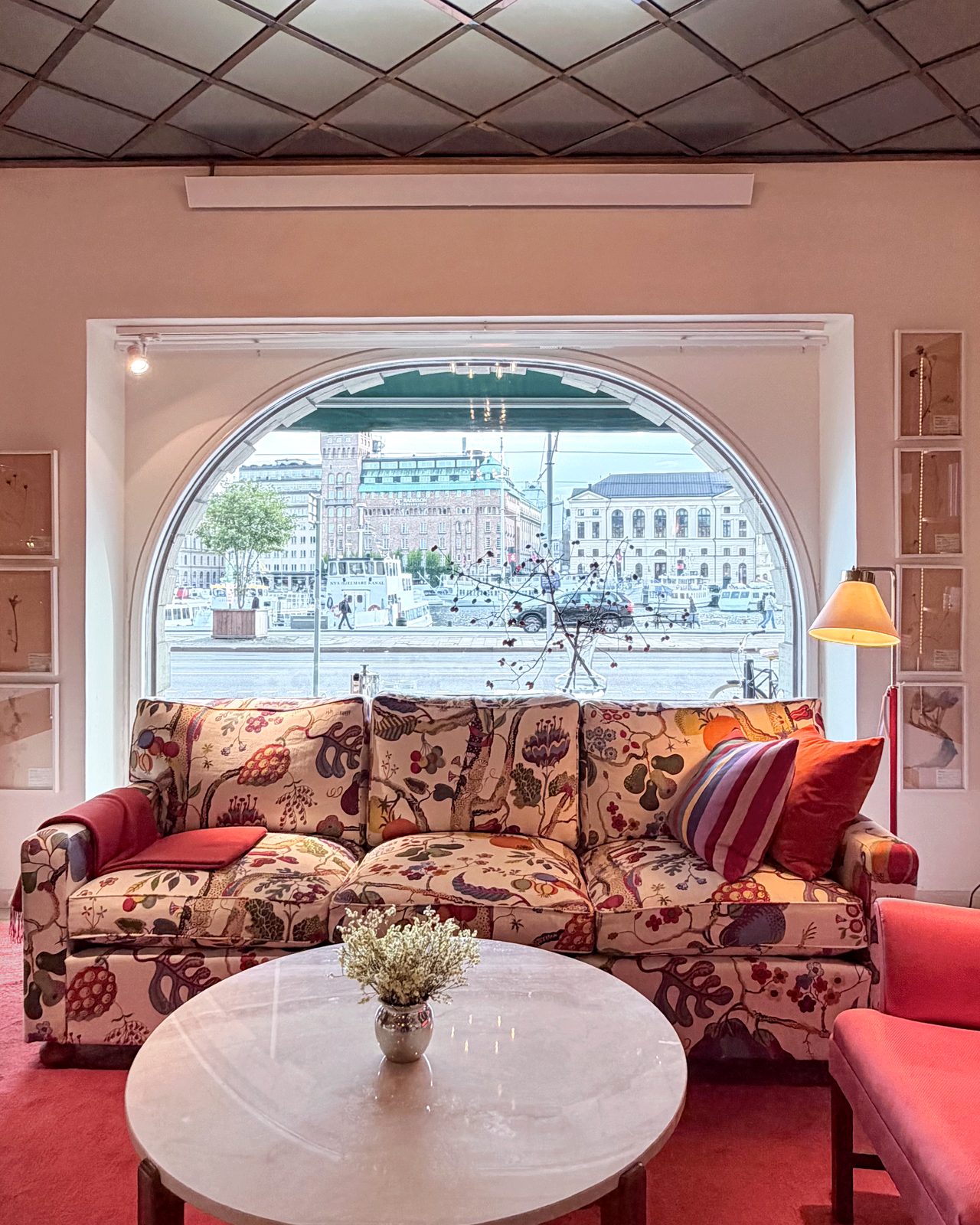 A sofa in a large floral print under an arched window looking out over the water in Stockholm, with a round marble table and a red carpet.