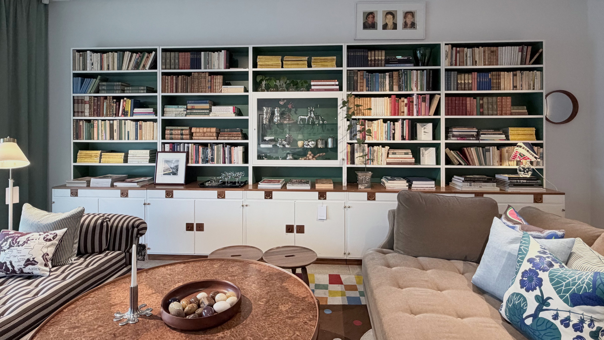 A floor to ceiling library wall above white cabinets, with a copper topped table and a bowl of stones in front.