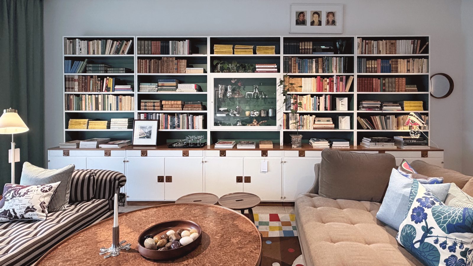 A floor to ceiling library wall above white cabinets, with a copper topped table and a bowl of stones in front.