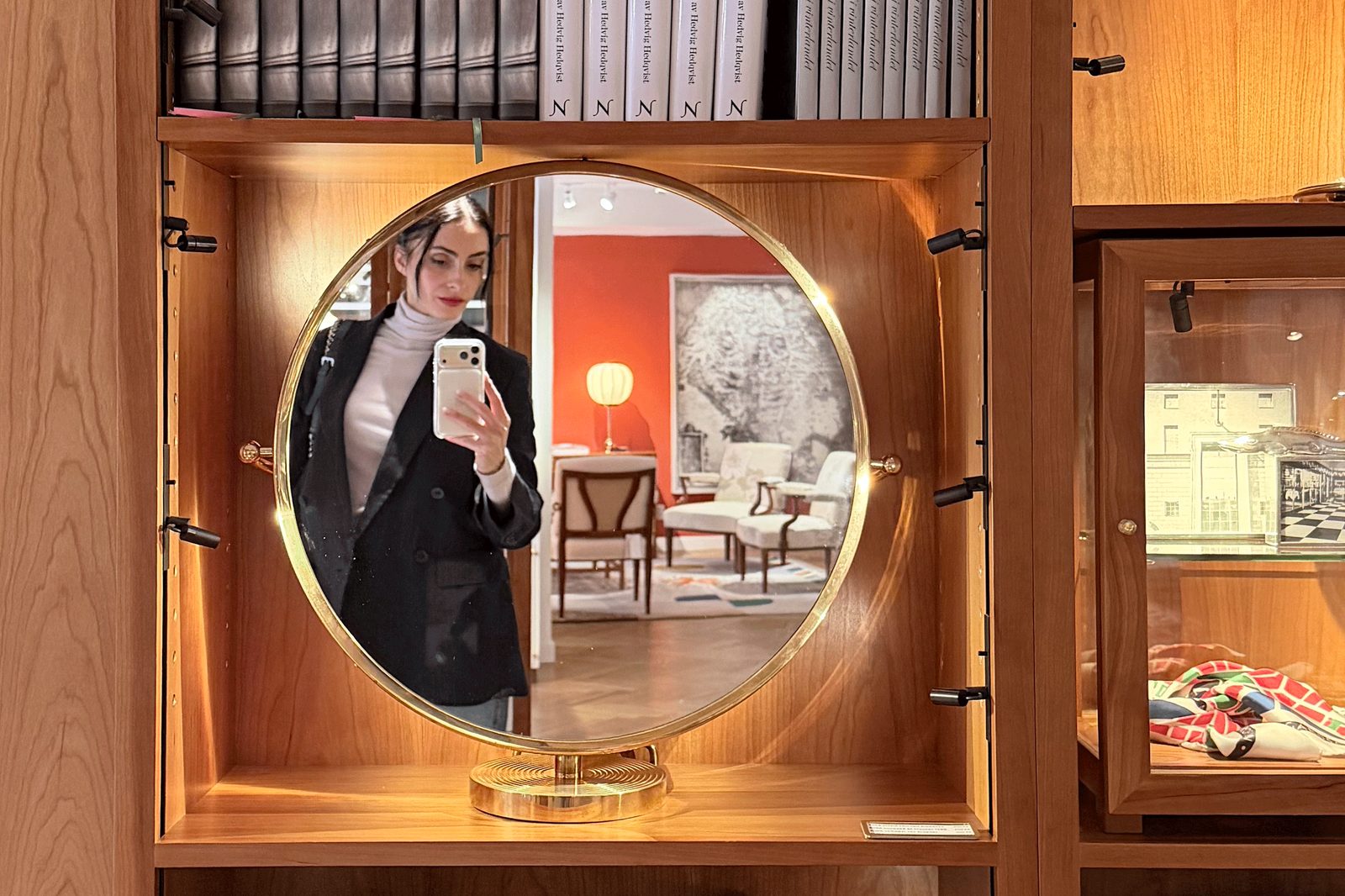 Madalina photographing herself in a round brass mirror inside a lit walnut display cabinet.