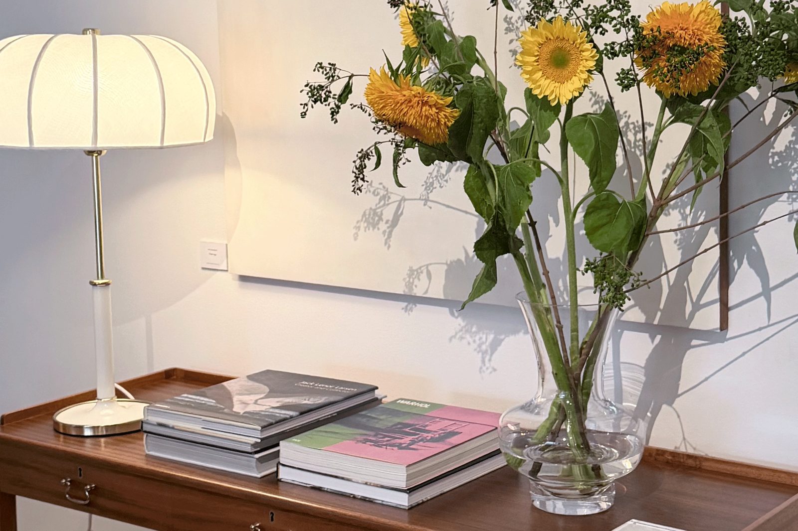 A walnut console against a pale wall with a white table lamp, a stack of books and a glass vase of sunflowers.