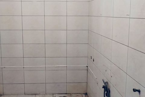 Bathroom before renovation: worn beige wall tile, exposed wiring, and an unfinished floor.